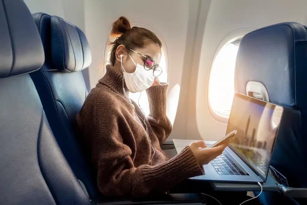 PassportCard/Internationale Krankenversicherung/woman-sitting-in-airplane-with-mask-looking-on-smartphone-with-laptop